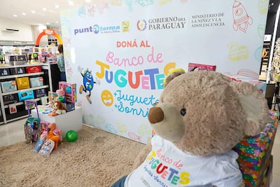 Un gran oso de peluche con camiseta del Banco de Juguetes rodeado de juguetes variados; ambiente de recolección en un local comercial.
