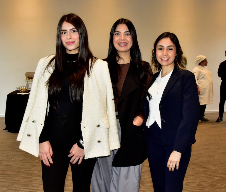 Tres mujeres sonrientes en primer plano: chaqueta blanca, abrigo negro y traje azul oscuro en un ambiente formal.