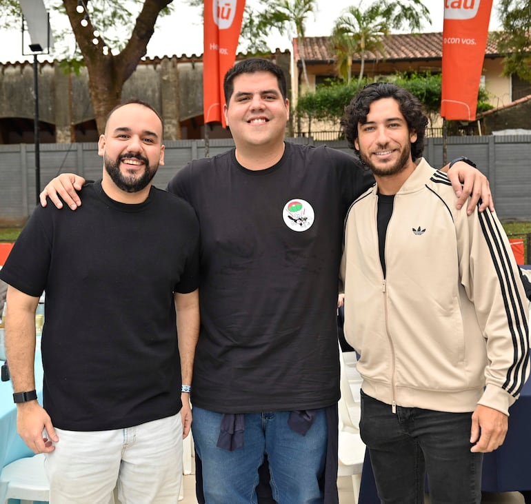 Tres hombres sonrientes, uno con barba y camiseta negra, otro con camiseta negra y emblema, y el tercero con chaqueta beige y cabello rizado.