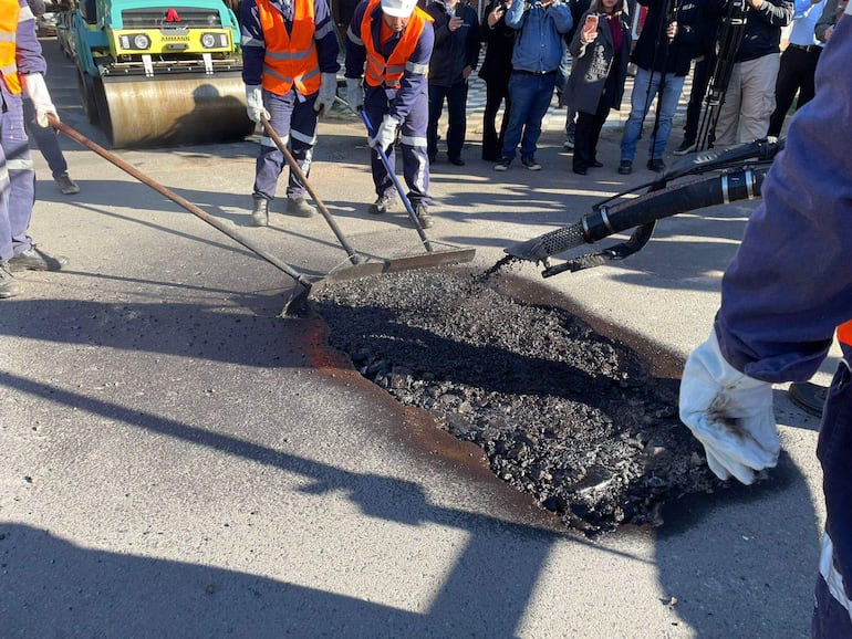 Cuatro operarios con chalecos naranjas y guantes utilizan palas y asfalto, mientras observadores registran la actividad con sus teléfonos.