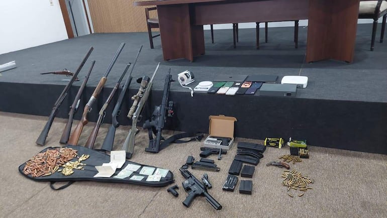 Rifles y pistolas sobre mesa gris, municiones y dinero en billetes, sin personas visibles en el entorno cerrado.