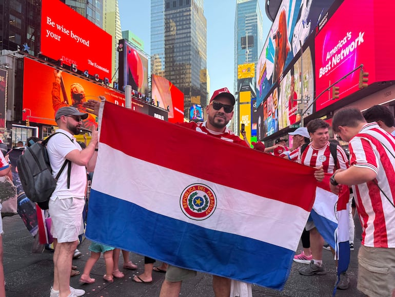 Hombre con camiseta roja y blanca sostiene la bandera de Paraguay en Times Square, rodeado de otros aficionados animados.