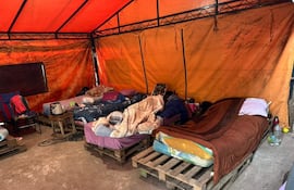 Camas improvisadas sobre pallets en albergue con familias abrigadas con mantas en carpas húmedas.