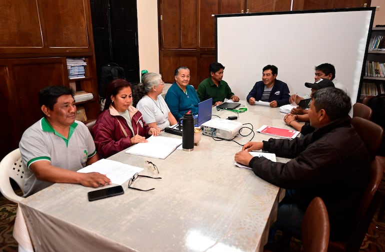 Ocho adultos, cuatro hombres y cuatro mujeres, discuten alrededor de una mesa, con una computadora y un proyector en el centro.