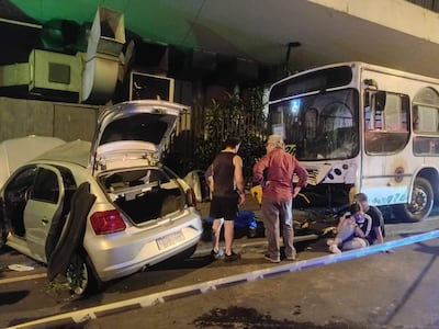 Mujer sentada en el suelo, con hombre mayor a su lado, mientras otros evalúan la situación cerca de un autobús dañado.