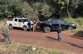 Cuatro personas, tres policías en uniforme y un civil, junto a camionetas en un camino rural con vegetación densa.