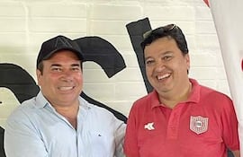 Dos hombres de pie, uno con gorra negra y camisa blanca, el otro con camiseta roja a rayas, se estrechan la mano.