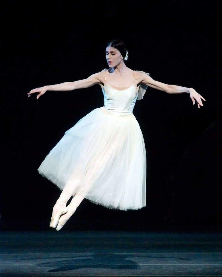 Bailarina en tutú blanco y zapatillas, decorada con flor blanca, ejecutando un elegante salto sobre fondo oscuro.