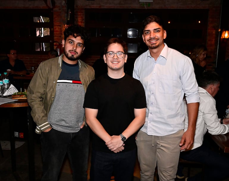Tres hombres sonrientes en un evento: uno con chaqueta verde, otro en camiseta negra, y el tercero en camisa blanca, rodeados de mesas.