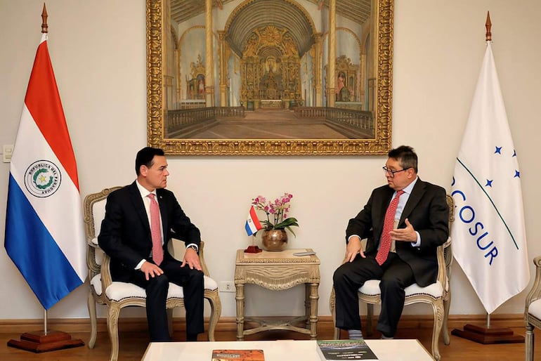 Rubén Ramírez Lezcano con corbata roja y Gustavo Alfredo Leite Gusinky en traje oscuro, conversando en un entorno formal con banderas.