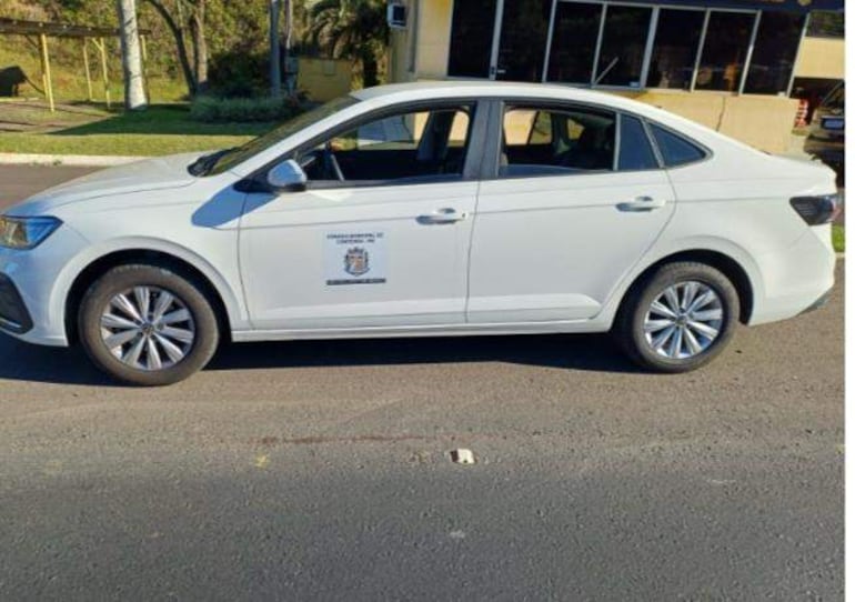 Coche sedán blanco estacionado junto a edificio con vegetación de fondo, sin personas visibles en la escena.