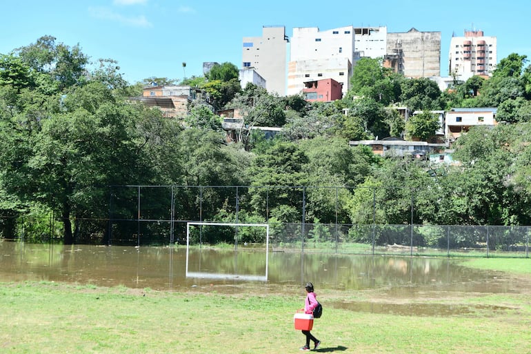 Persona con chaqueta rosa y cooler rojo camina en campo de fútbol lleno de agua, rodeado de edificios deteriorados.