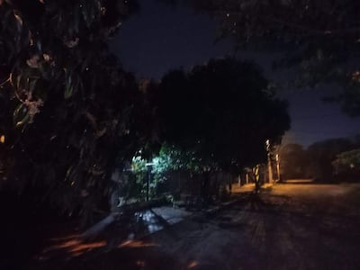 Calle vacía y oscura en Villa del Maestro, iluminada por un farol, rodeada de árboles.