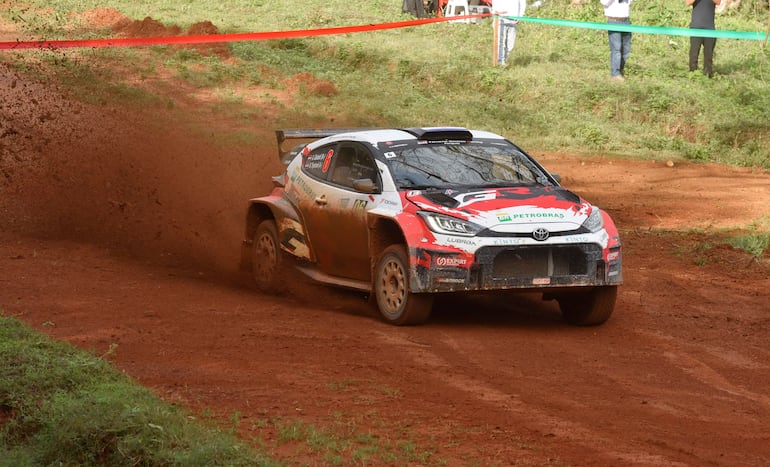 Automóvil de rally Toyota salpicando tierra en un camino, con espectadores observando en el fondo.