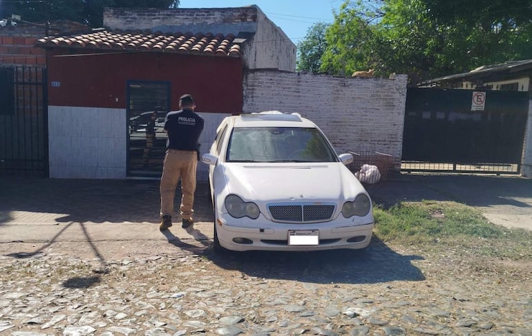 Oficial de policía de espaldas en uniforme negro observa un vehículo blanco. Área urbana con cerca de metal y gato en la pared.