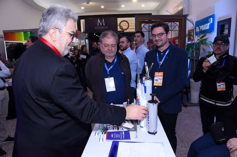 Hombre de cabello gris con traje oscuro y gafas interactúa en una mesa con productos, rodeado de otros expositores atentos.