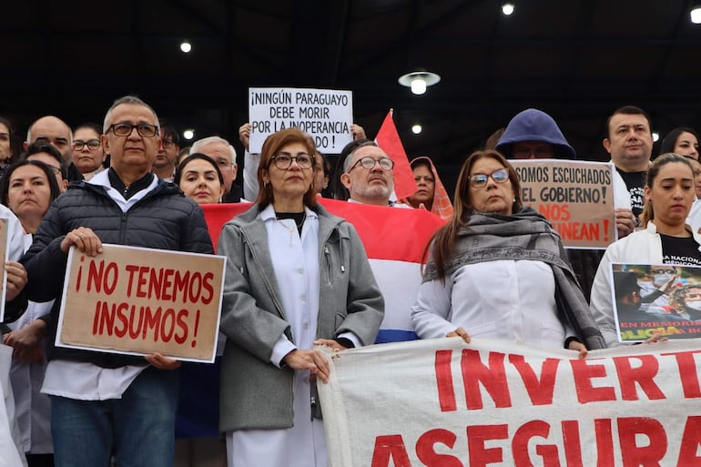 Marlim Riveros Pesoa y Syrley Ferreira sostienen carteles en una manifestación, vistiendo batas blancas, rodeados de banderas paraguayas.