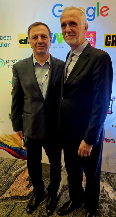 Dos hombres de pie en un evento, uno con chaqueta azul y otro en traje negro, ambos con expresiones serias ante logotipos corporativos en el fondo.