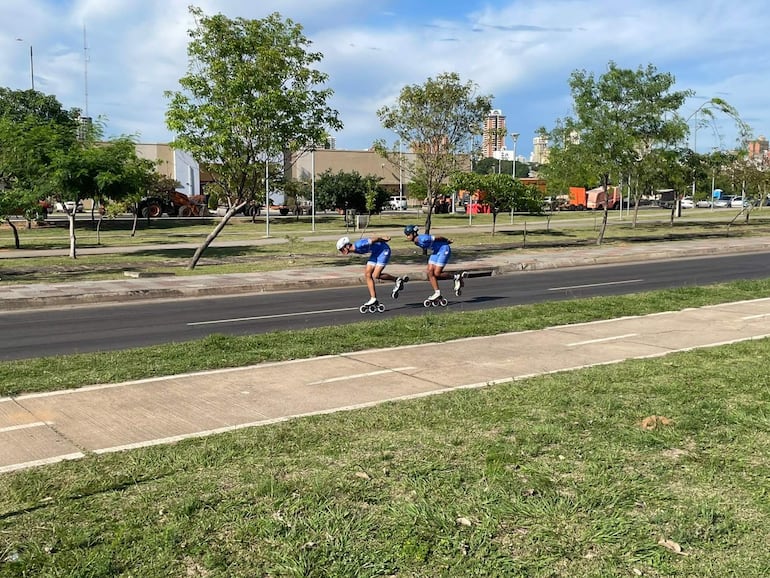Dos patinadores en camisetas azules y pantalones cortos, rodeados de árboles y un ambiente urbano soleado.