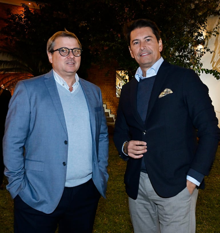 Dos hombres conversan, uno con gafas y blazer claro, el otro con blazer oscuro, en un evento al aire libre con césped y ladrillo al fondo.
