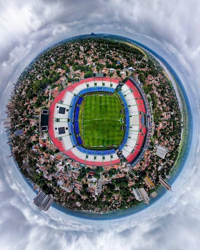 Estadio Defensores del Chaco con gradas rojo, blanco y azul. Campo de juego bien mantenido y cielo parcialmente nublado.