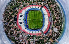 Estadio Defensores del Chaco con gradas rojo, blanco y azul. Campo de juego bien mantenido y cielo parcialmente nublado.