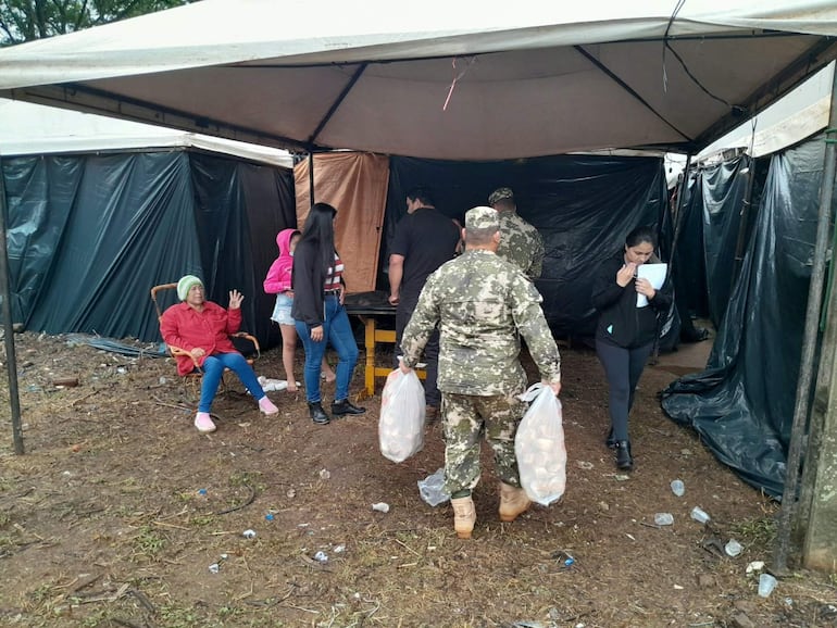 Mujer mayor con abrigo rojo sentada, joven escucha, hombre militar camina con bolsas hacia las carpas en un refugio desordenado.