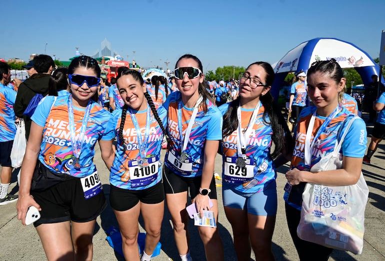 Cinco mujeres sonrientes en camisetas azules y naranjas, preparándose para la carrera en un entorno exterior despejado.