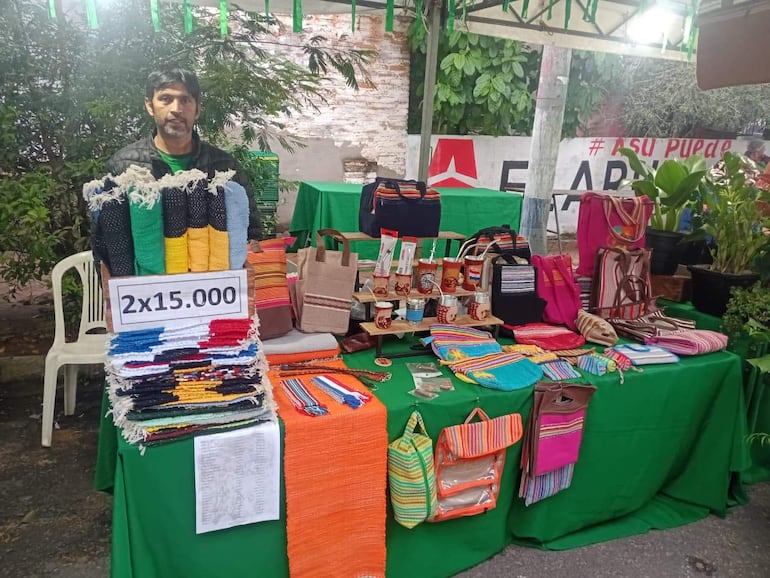 Vendedor de artesanías con cabello largo y abrigo oscuro, de pie tras mesa cargada de coloridas piezas de tela y souvenirs.