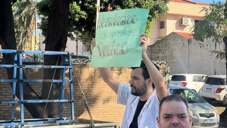 Un hombre en camisa blanca sostiene un cartel que dice 'Atendemos con las manos Vacías' mientras otro lo observa seriamente.