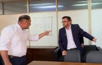 Guido Benítez en camisa blanca señala a Maximiliano Seiferheld en chaqueta oscura, en oficina con pizarra blanca al fondo.