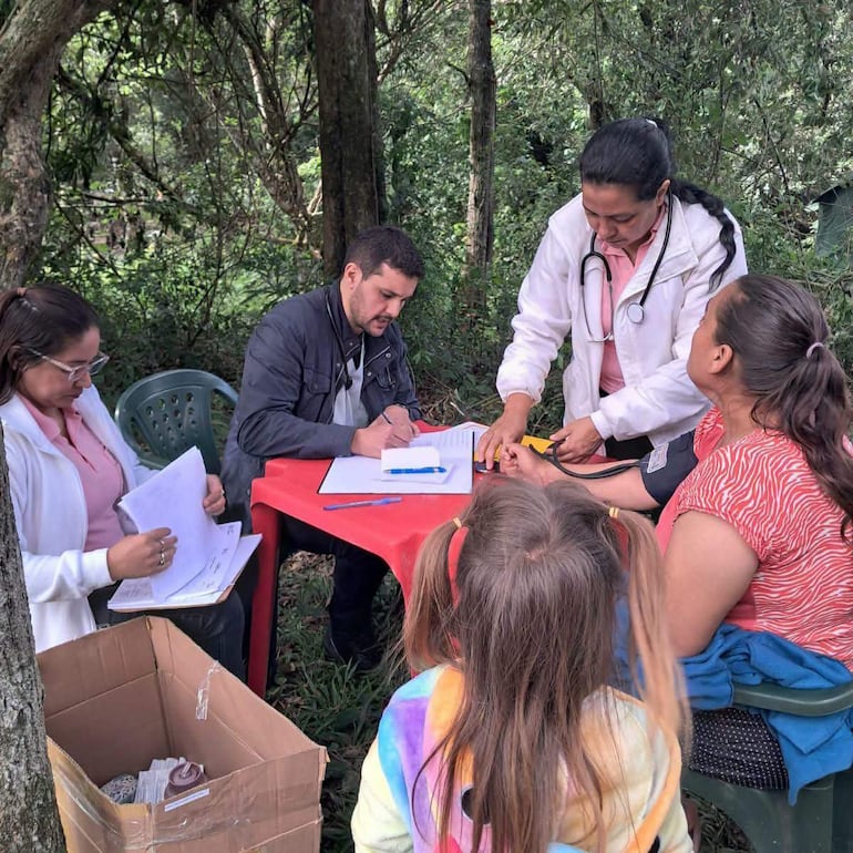 Hombre con chaqueta escribe en mesa roja, mientras mujeres asisten a pacientes en un entorno natural, un ambiente de atención médica comunitaria.
