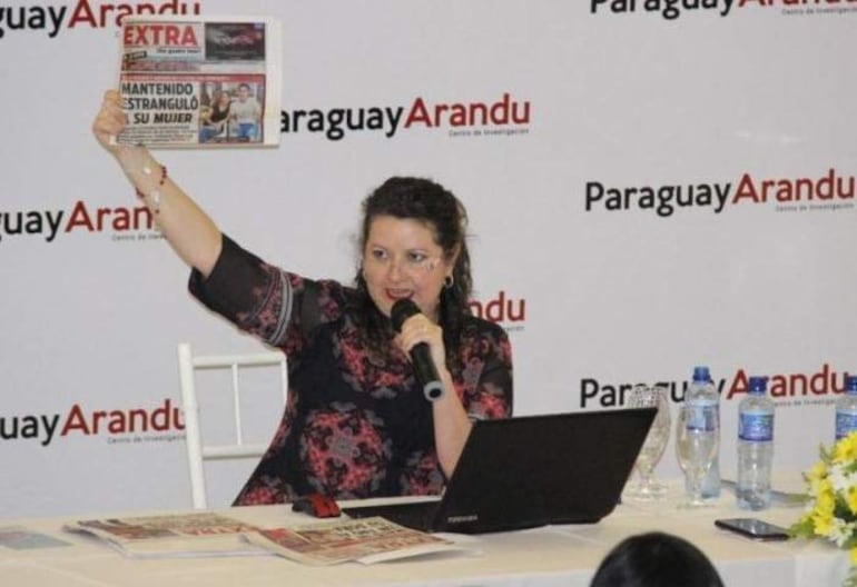 Lidia Mariana Fariña Sanabria con blusa negra y detalles florales, sostiene un periódico y un micrófono frente a una audiencia.