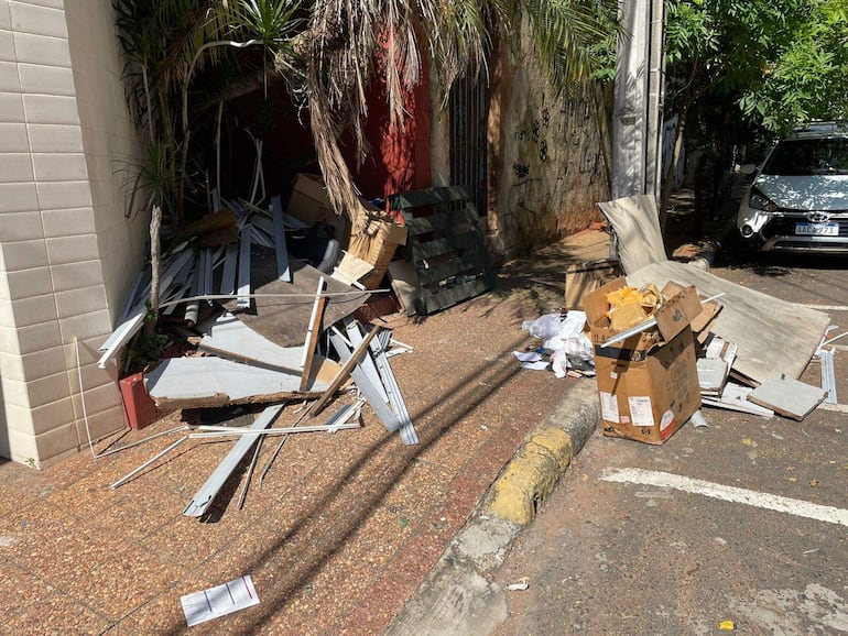 Espacio exterior con desechos amontonados y coche estacionado al fondo, en un área urbana con vegetación.