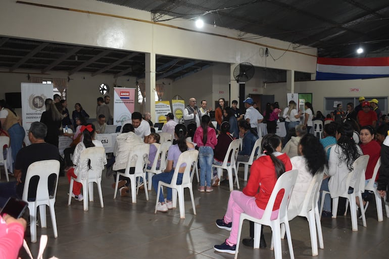 Grupo de personas conversando en un salón, con niños y adultos, y una bandera de Paraguay al fondo.
