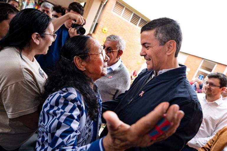 Santiago Peña sonríe y estrecha la mano de una mujer con chaqueta distintiva, mientras otros observan en un ambiente cálido.