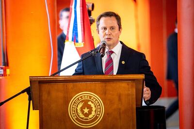 Alberto Joaquín Martínez Simón en traje oscuro y corbata a rayas, habla frente a un micrófono con una bandera de Paraguay de fondo.