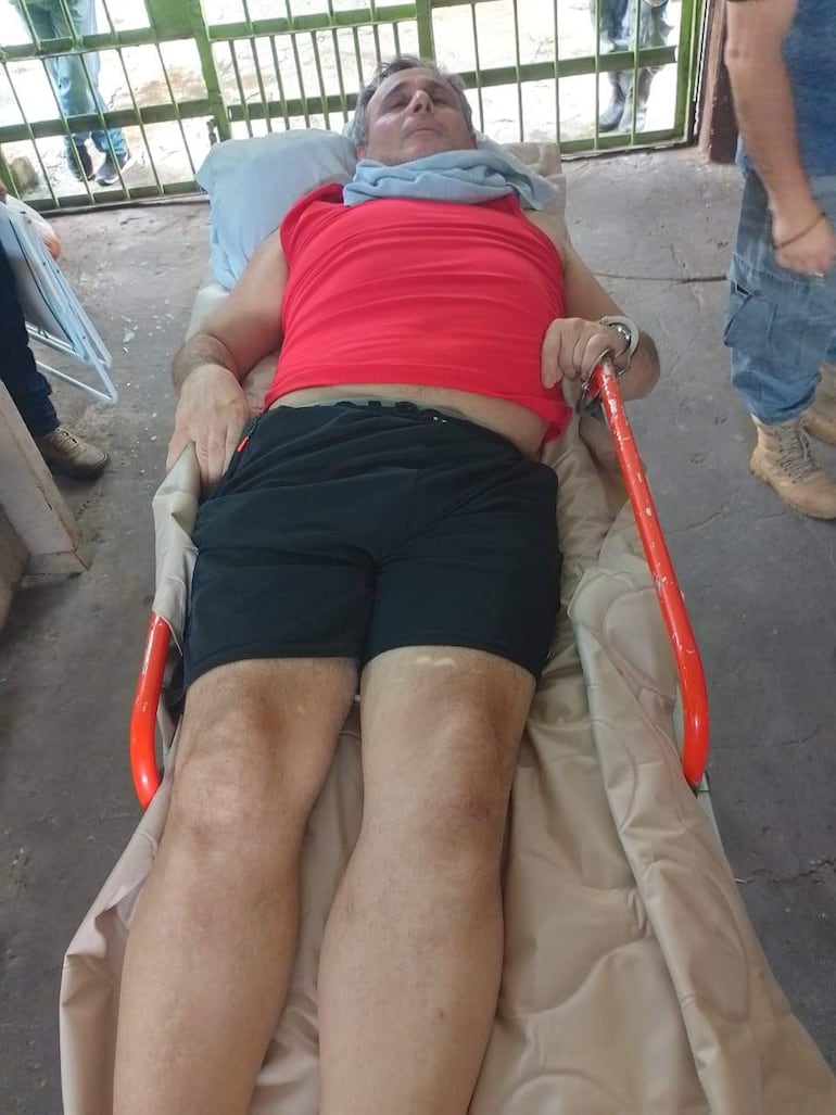Acosta Marques recostado en una camilla, con camiseta roja y pantalones cortos negros, en un ambiente médico desorganizado.