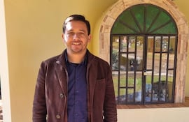 Ariel Girett en chaqueta marrón y camisa azul, sonriente, frente a una ventana en un entorno exterior.