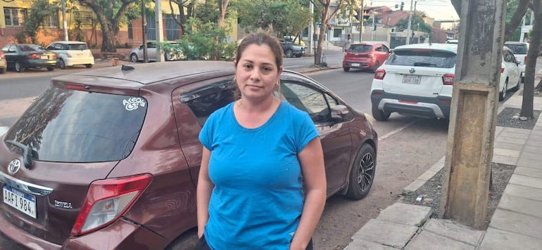 Fáitima Patiño de pie junto a un automóvil marrón, con camiseta azul y expresión seria en una calle urbana.
