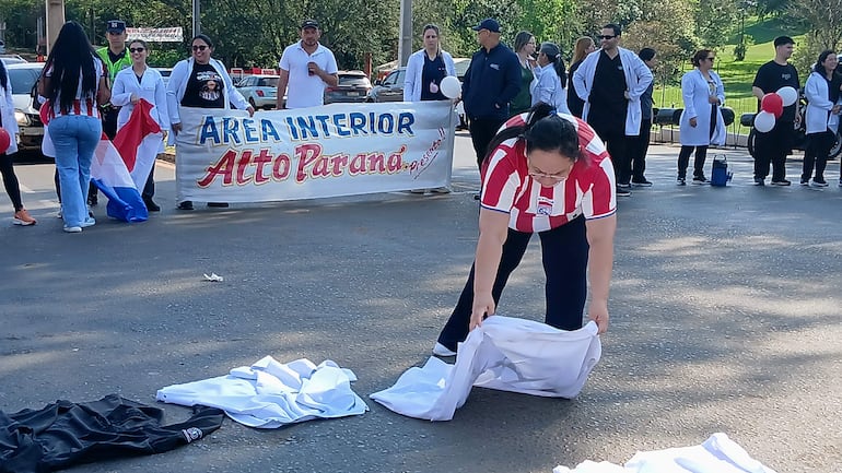 Mujer con camiseta a rayas rojas y blancas organiza objetos en manifestación, rodeada de médicos con batas blancas y globos rojos y blancos.