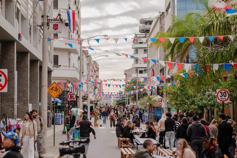 Ambiente urbano festivo con banderas rojo, blanco y azul; personas interactuando en mesas al aire libre.