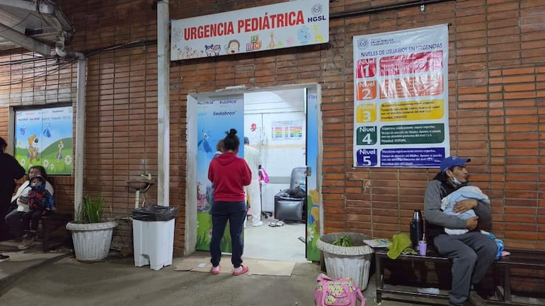 Mujer de abrigo rojo y niño, junto a un hombre con un bebé y una botella, en ambiente hospitalario con carteles sobre urgencias.
