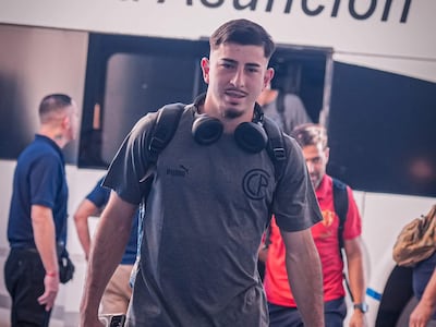 Joven con camiseta gris y auriculares, sosteniendo botella y maleta, rodeado de otros con camisetas rojas en un ambiente informal.