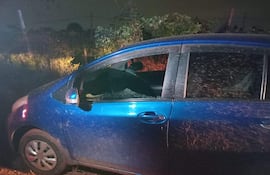 Automóvil azul sucio y desgastado con ventana rota, estacionado en un entorno rural y rodeado de vegetación.