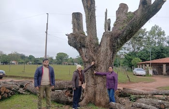 Hombre robusto con chaqueta azul y dos mujeres con abrigos, observan un tajy antiguo rodeado de vegetación y troncos apilados.