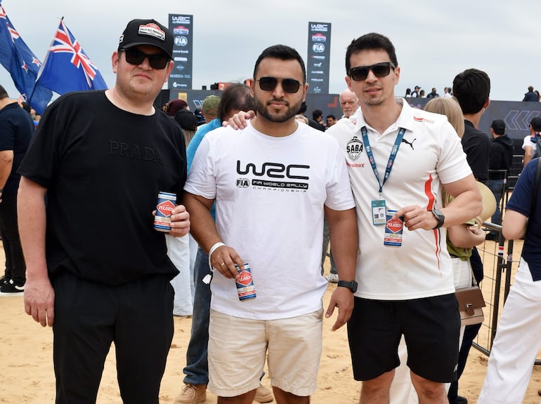 Tres hombres con gafas de sol, uno en camiseta negra de Prada y otros en blancas del WRC, bebiendo cerveza en evento de rally.