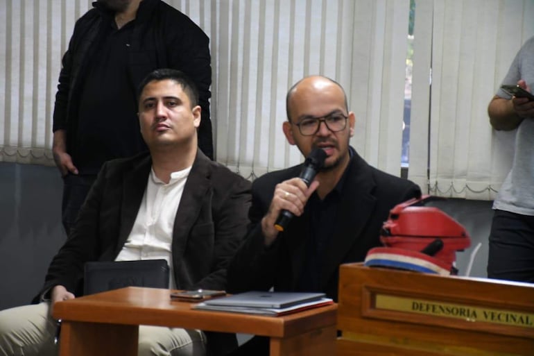 Hombre con gafas y micrófono hablando, mientras otro escucha pensativo en una oficina con documentos sobre la mesa.
