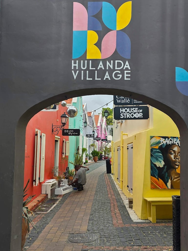 Hombre de espaldas agachado junto a una fuente en HULANDA VILLAGE, rodeado de edificios coloridos.