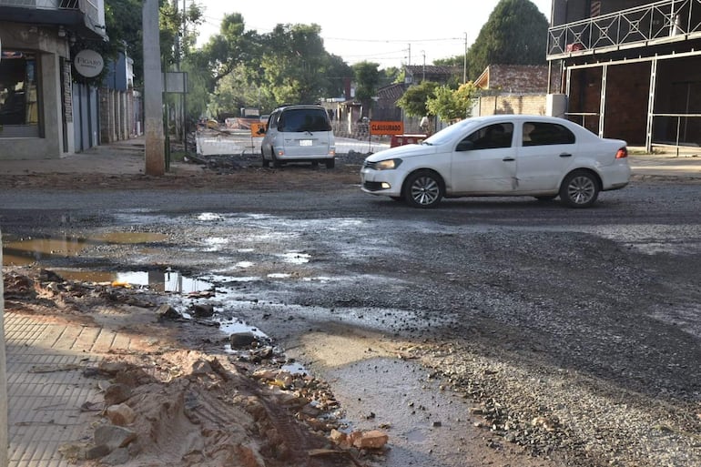 Escena urbana con un automóvil blanco en movimiento y un vehículo gris estacionado, rodeados de baches y charcos de agua.